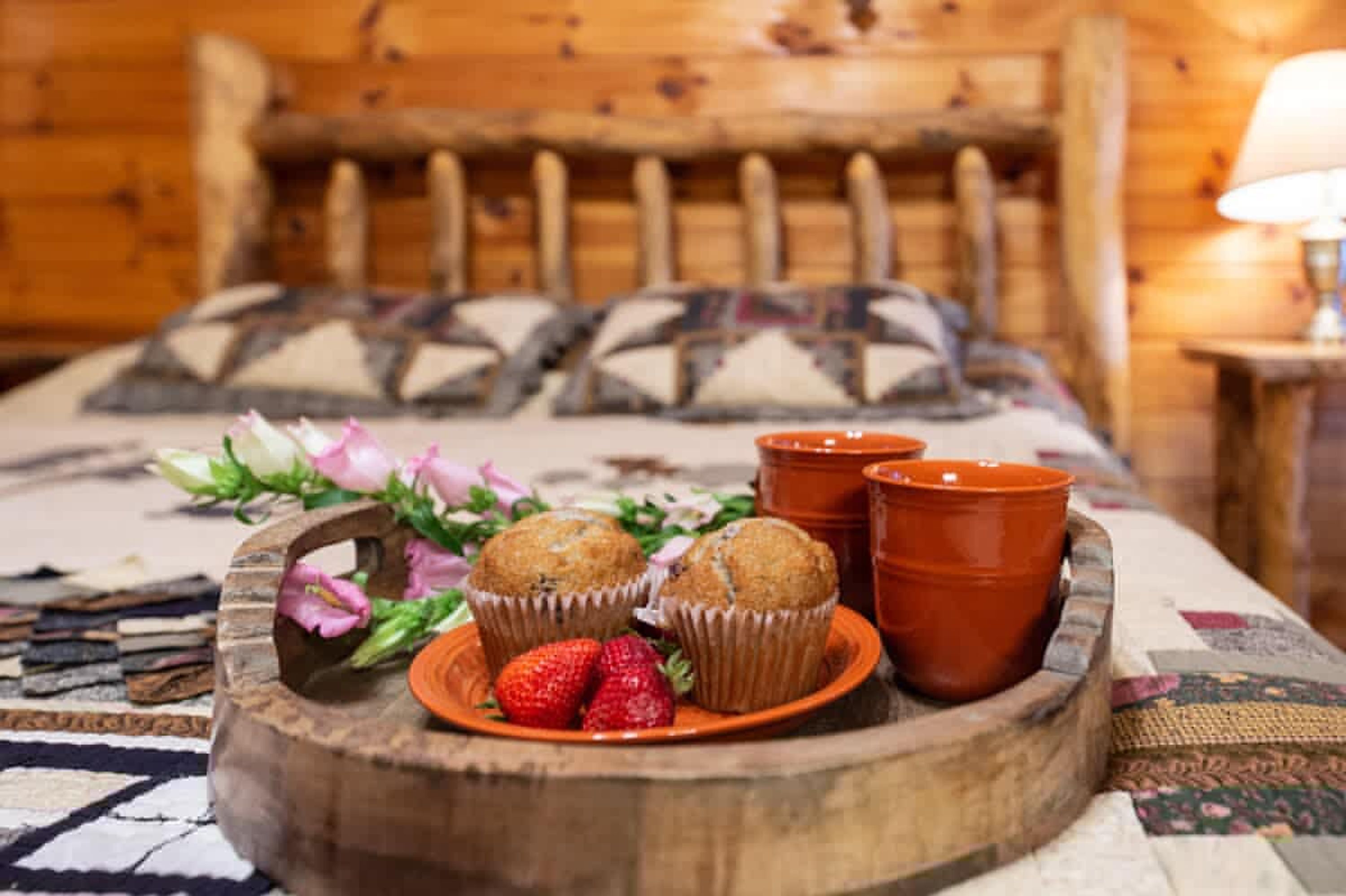 A wooden bed with patterned pillows, a tray with muffins and strawberries, and two terracotta cups.