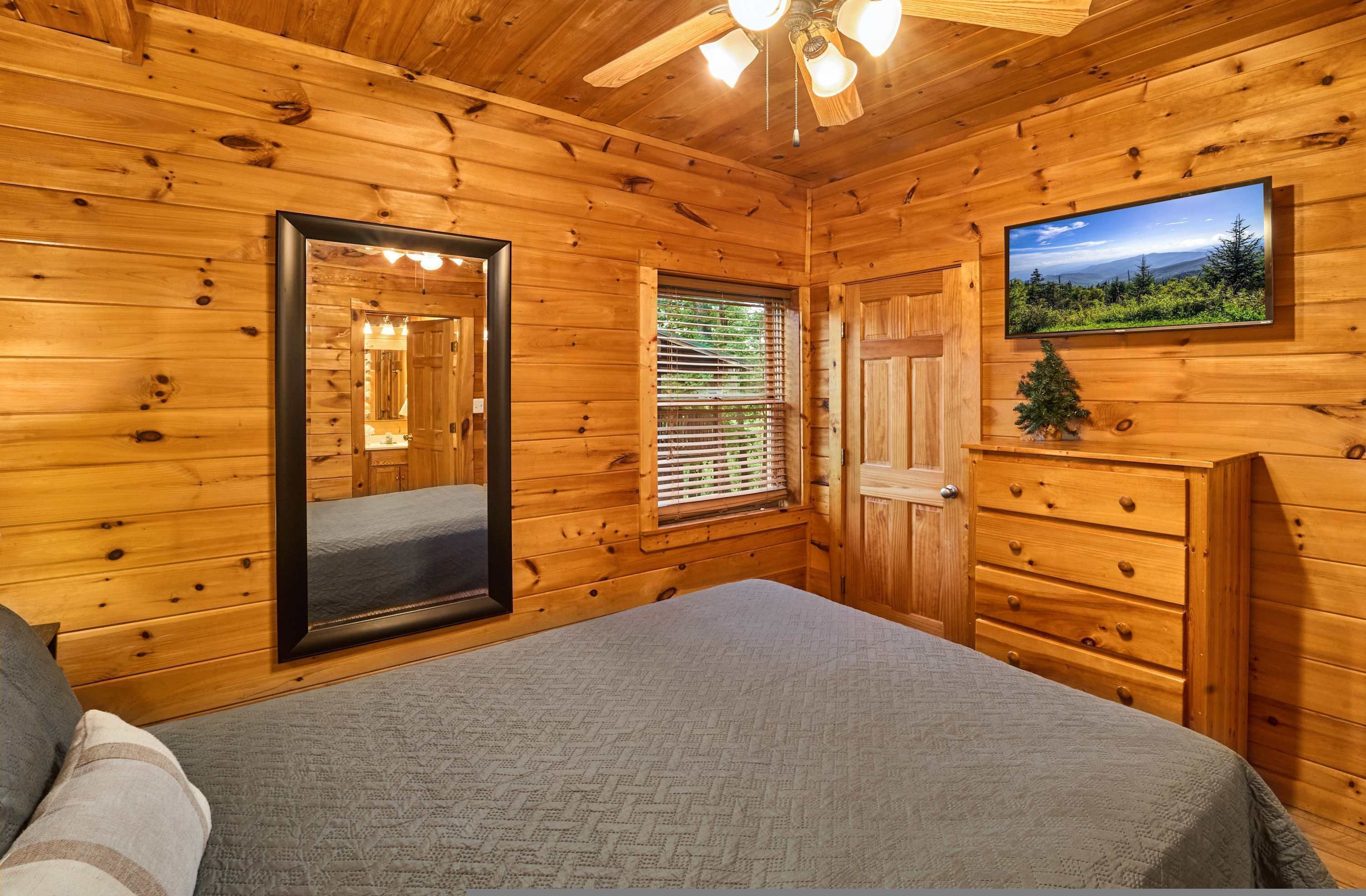 Une chambre à coucher dans un chalet en bois, avec un lit, une commode, un miroir, une fenêtre avec des stores et un téléviseur fixé au mur.
