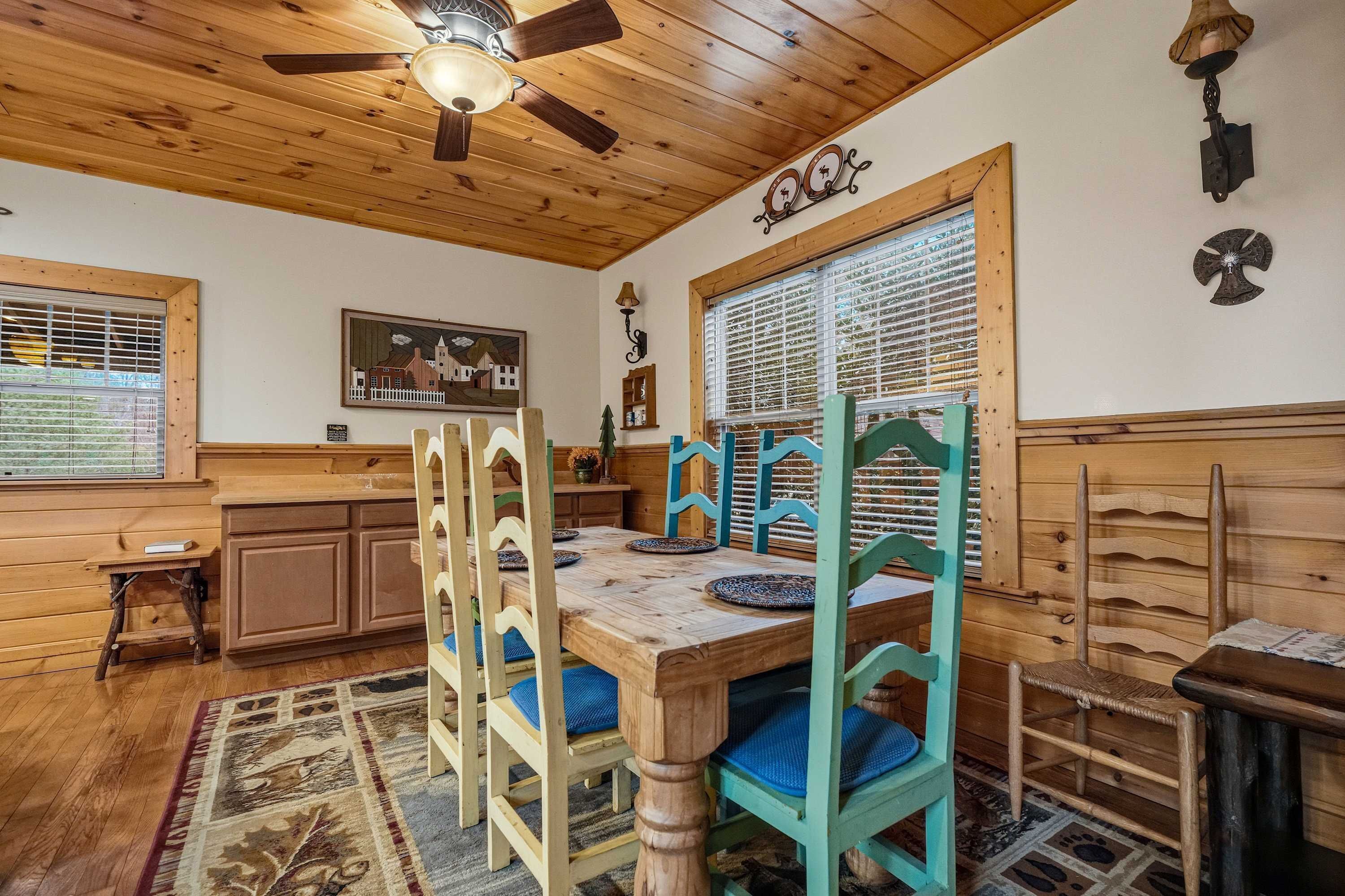 A wooden dining area with a table set for four, wooden chairs, a ceiling fan, and a window with blinds.
