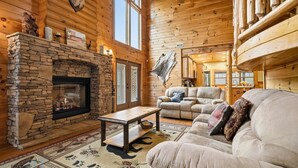 A log cabin interior with a stone fireplace, leather sofa, and a large window. - 5BR Cabin w Yard Office Hot Tub Firepit (Sevierville)