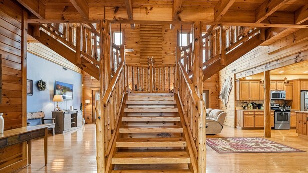 A wooden staircase leading to a second floor, flanked by wooden railings, with a kitchen and living area visible on the landing. - 5BR Cabin w Yard Office Hot Tub Firepit (Sevierville)