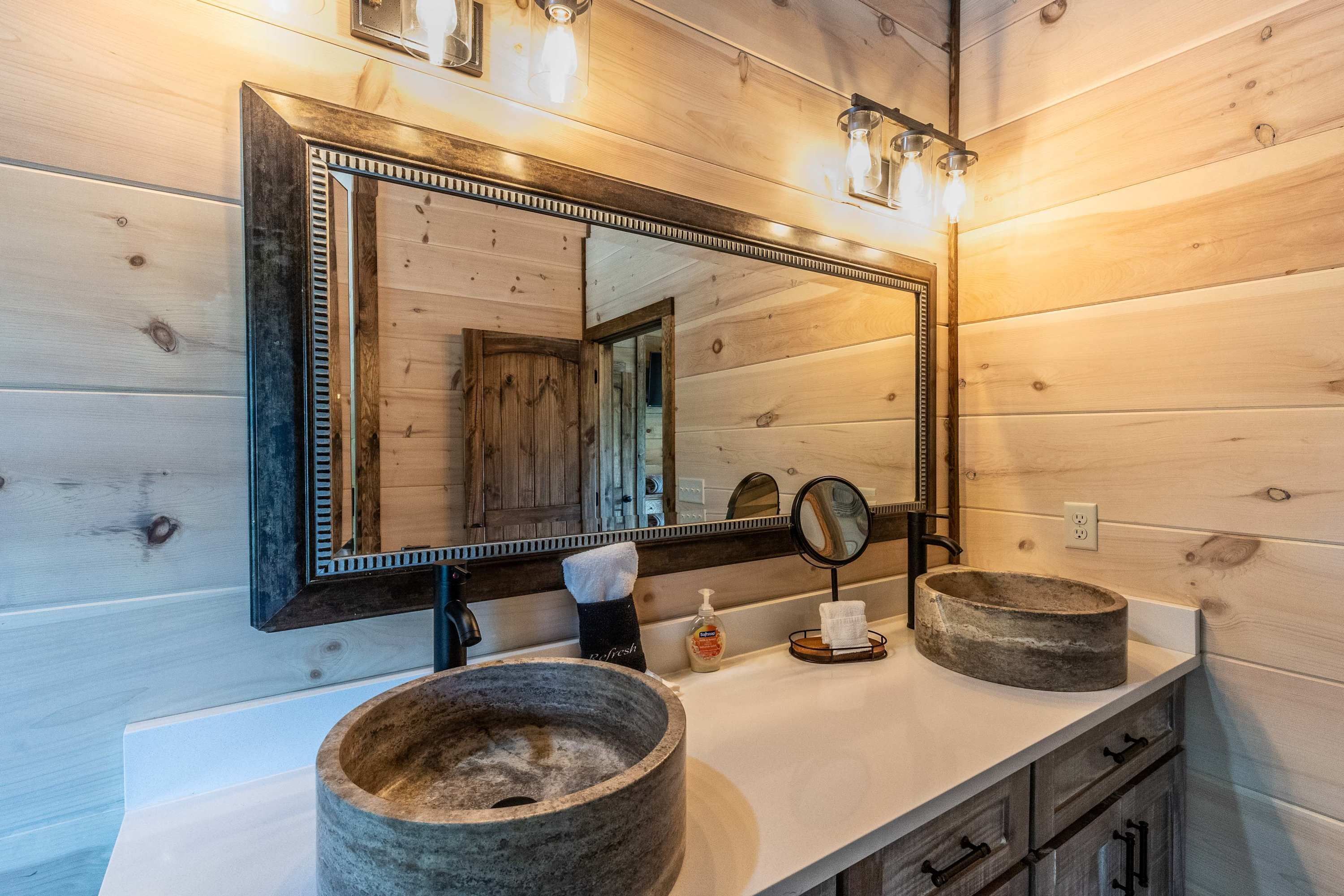 A bathroom with wooden panel walls, a large mirror, two stone basins, a towel rack, and a soap dispenser.