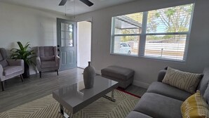 Un salon avec un canapé gris, deux fauteuils, une table basse et une grande fenêtre équipée de stores.