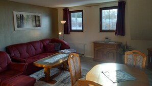 Una sala de estar con un sofá rojo, una mesa de centro de madera, una mesa de comedor con sillas, un radiador y una ventana con cortinas.