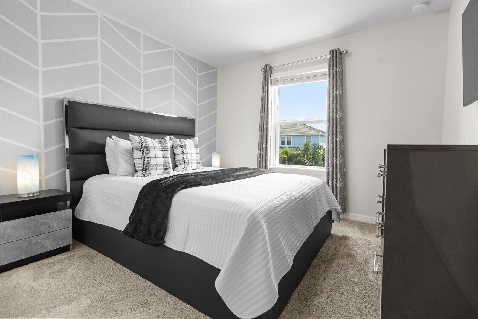A bedroom with a bed, nightstands, a dresser, a window with curtains, and a view of a building outside.