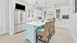 Una cucina moderna con mobili bianchi, un frigorifero in acciaio inossidabile, un'isola integrata con un tocco di azzurro, sedie in vimini, illuminazione a sospensione e un'ampia finestra.