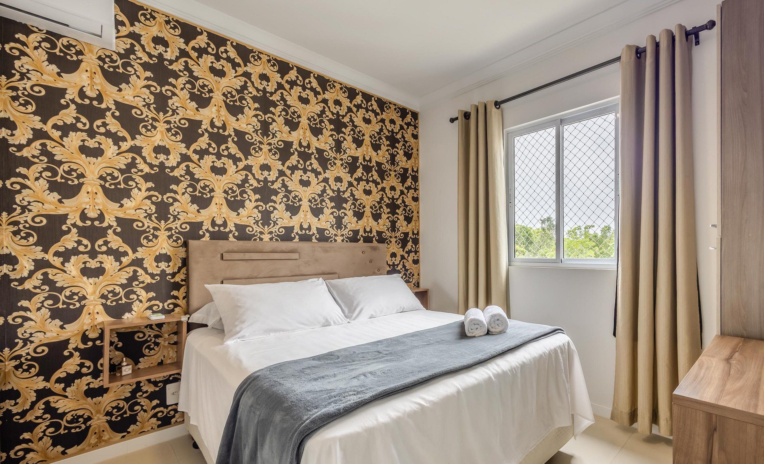 A bedroom with a bed, a nightstand, a window with curtains, and a wall with a patterned wallpaper.