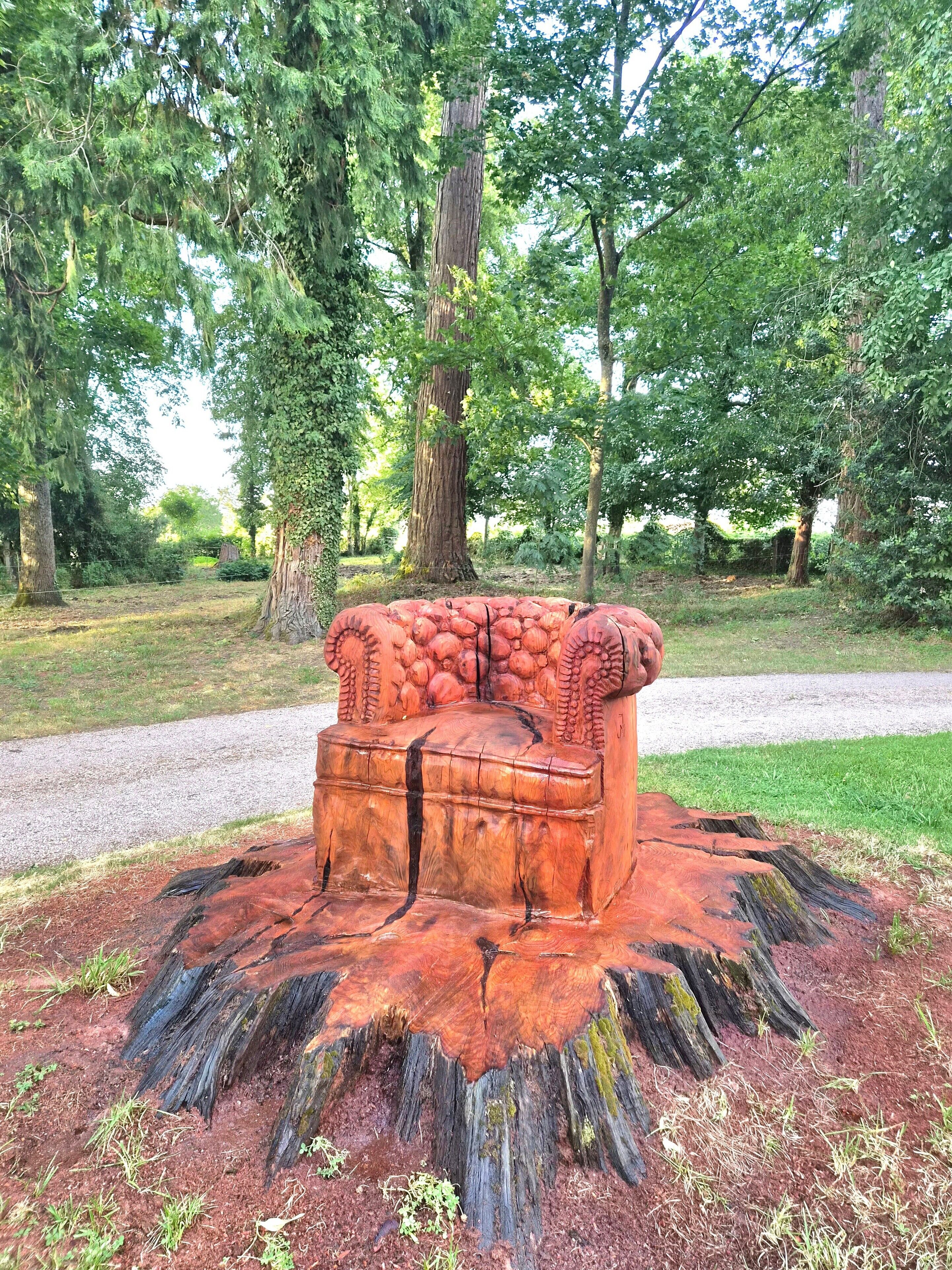 A wooden bench with carved details, surrounded by a tree stump base, set in a park with trees and a gravel path.
