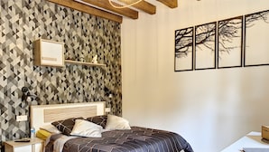 Un dormitorio con pared de diseño geométrico, cama con colcha estampada oscura, mesitas de noche con lámparas y un pequeño estante con adornos.