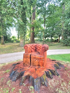 Un banco de madera con detalles tallados, rodeado por una base de tocón de árbol, ubicado en un parque con árboles y un sendero de grava.