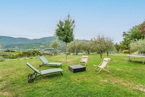 Un'area esterna erbosa con poltrone relax e un tavolo, circondata da alberi e colline.