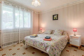 Un dormitorio con una cama, dos mesitas de noche, un ventanal con cortinas de encaje, un radiador y un cuadro colgado en la pared.