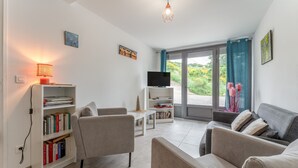 Een moderne woonkamer met een bank, fauteuil, boekenkast, televisie en uitzicht door een glazen deur.