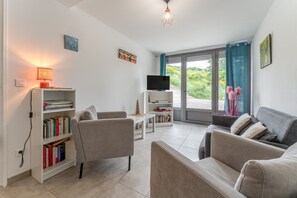 A modern living room with a sofa, armchair, bookshelf, television, and a view through a glass door. - Gîte Des Lavandes Apartment with Mountain View and Wi-Fi (Nyons)