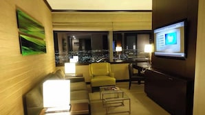 Une chambre d’hôtel moderne avec vue sur la ville, comprenant un canapé, un fauteuil jaune, une table basse en verre et un téléviseur à écran plat fixé au mur.