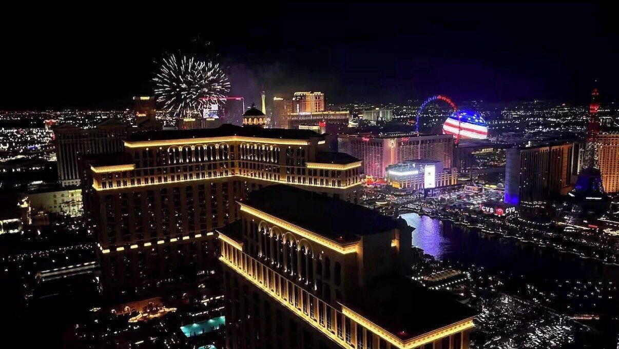 A nighttime cityscape with illuminated buildings, including a prominent hotel with a classic facade and a large fountain display.