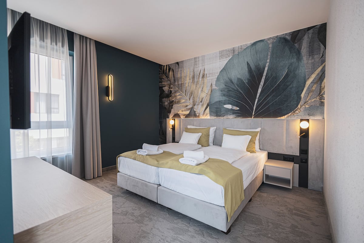 A modern hotel room with a large bed, a bedside table, a wall-mounted light, and a large leaf artwork.
