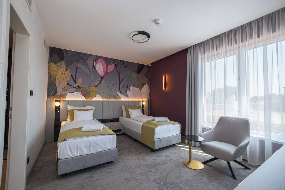 A hotel room with two beds, a chair, a nightstand, a lamp, and a large window with curtains.