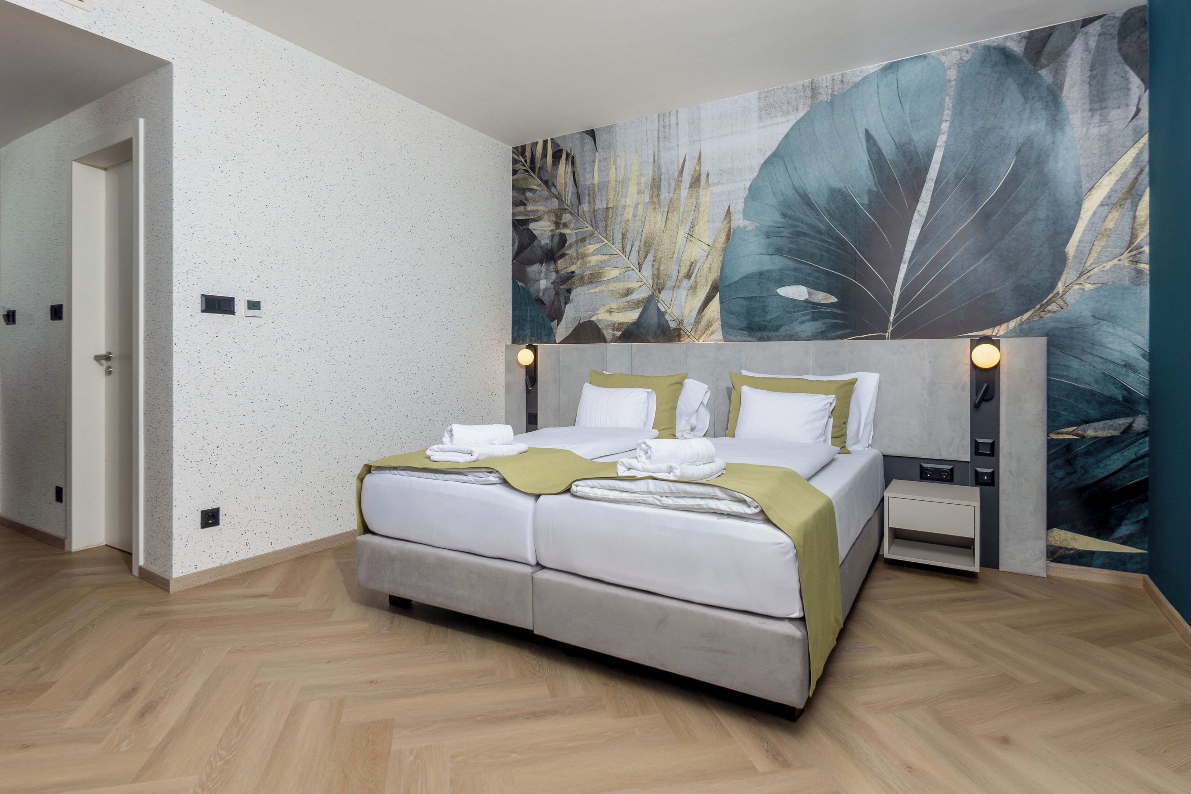 A modern hotel room with a large bed, a nightstand, a wall-mounted lamp, and a door leading to another room.
