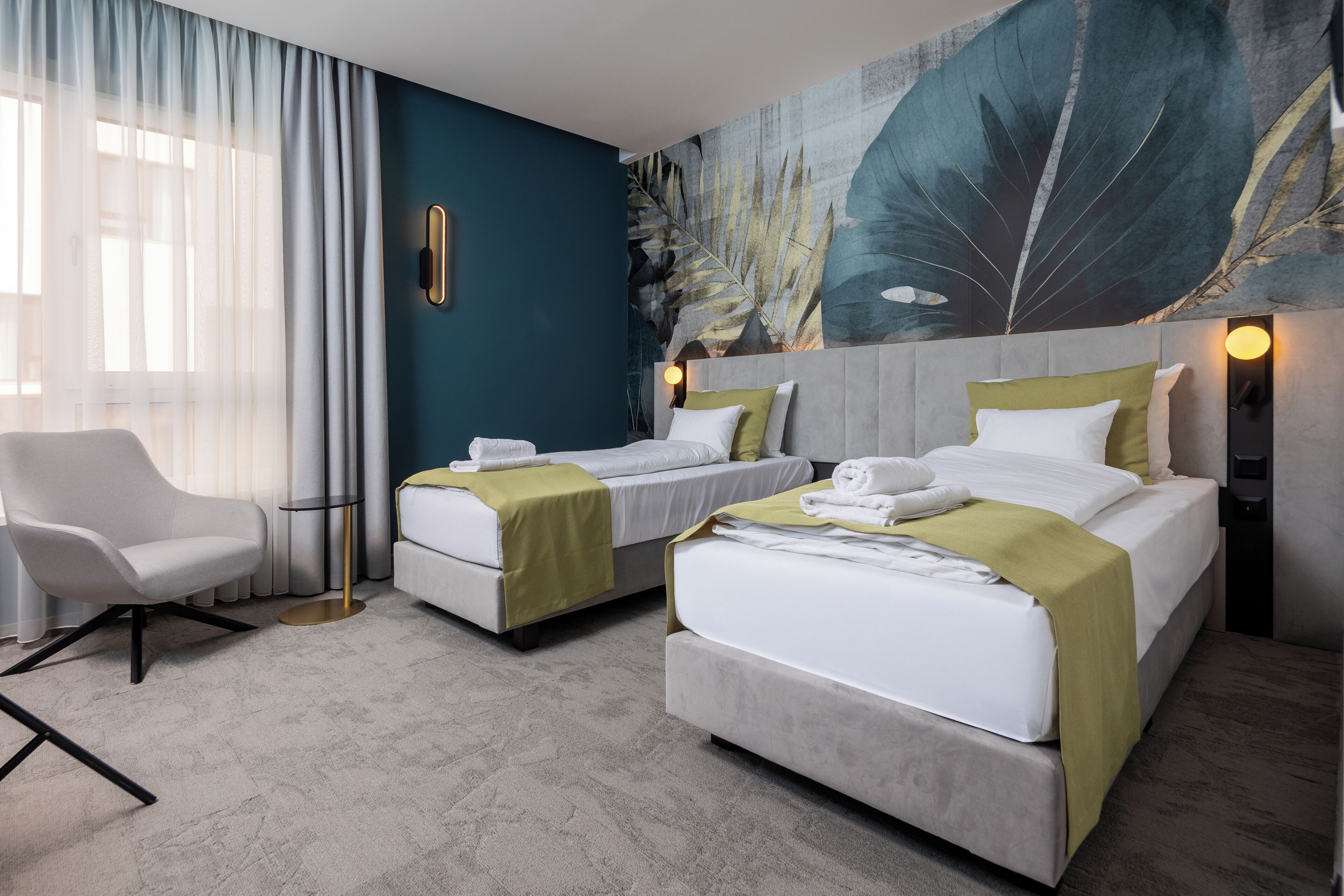 A hotel room with two beds, a chair, a small table, and a large leaf-patterned wall.