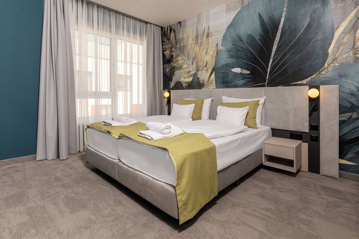 A double bed with a grey headboard, a bedside table, a wall-mounted light, and a large leaf-patterned mural.