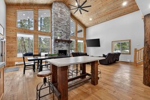 A spacious room with a stone fireplace, wooden ceiling, large windows, and a flat-screen TV.