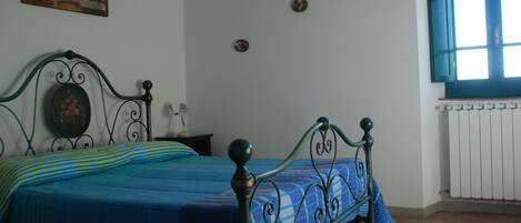 Un dormitorio con estructura de cama de metal, colcha azul y verde a rayas, un cuadro en la pared y una ventana con contraventanas cerradas.