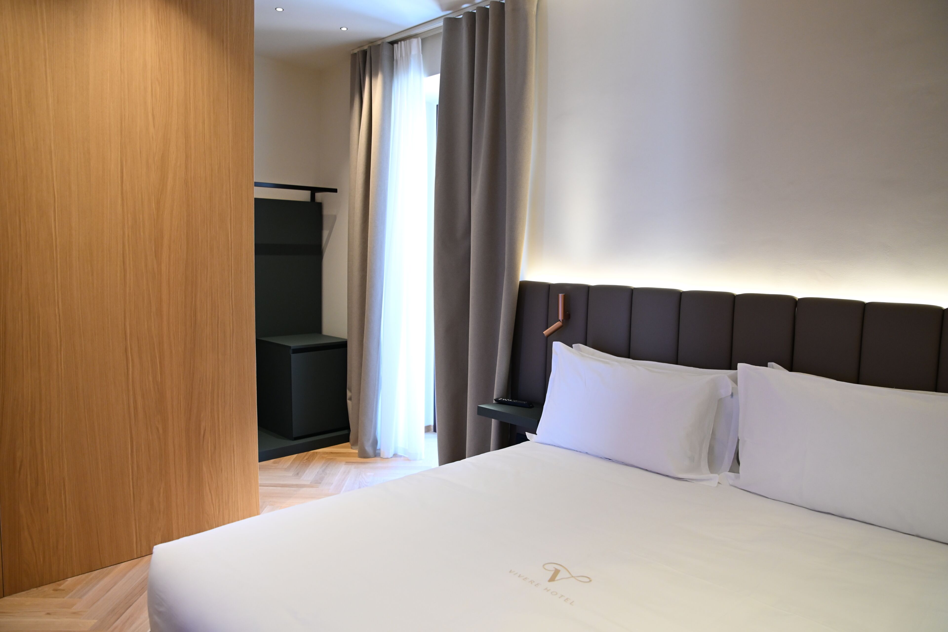 Habitación de hotel con una cama grande, un armario de madera, una mesita de noche oscura y vistas a una ventana con cortinas.