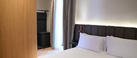 Habitación de hotel con una cama grande, un armario de madera, una mesita de noche oscura y vistas a una ventana con cortinas.