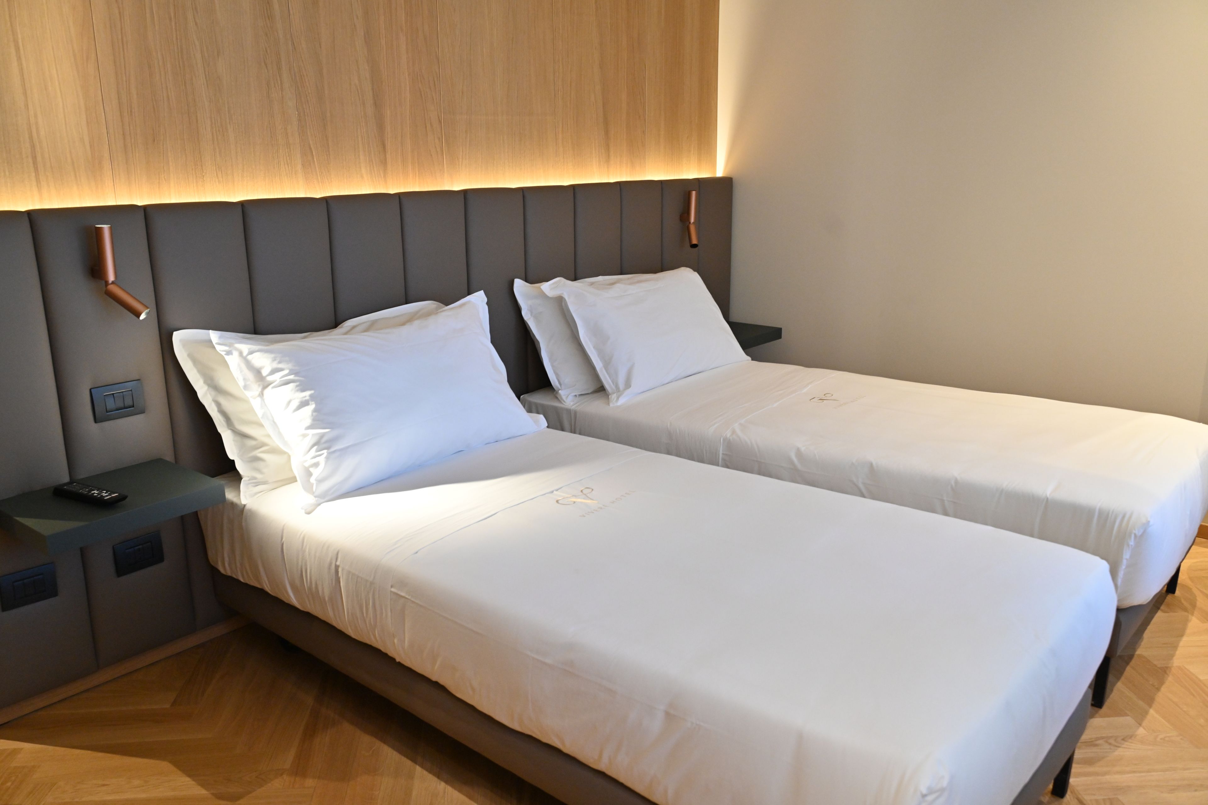Una camera d'albergo con due letti singoli, una testiera in legno, comodini e un tavolino con un telecomando.