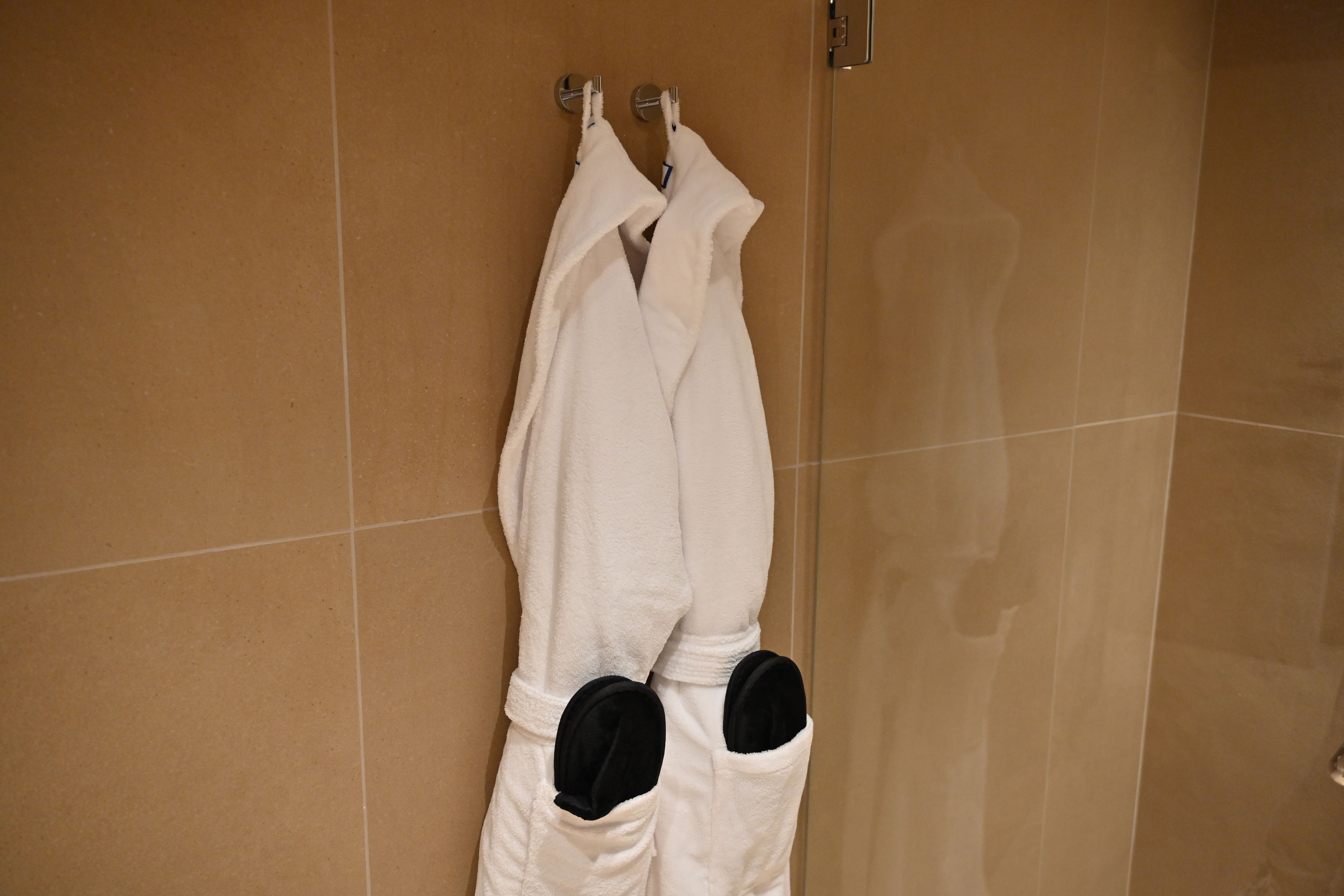 A white bathrobe hanging on a metal hook in a bathroom with beige tiled walls.