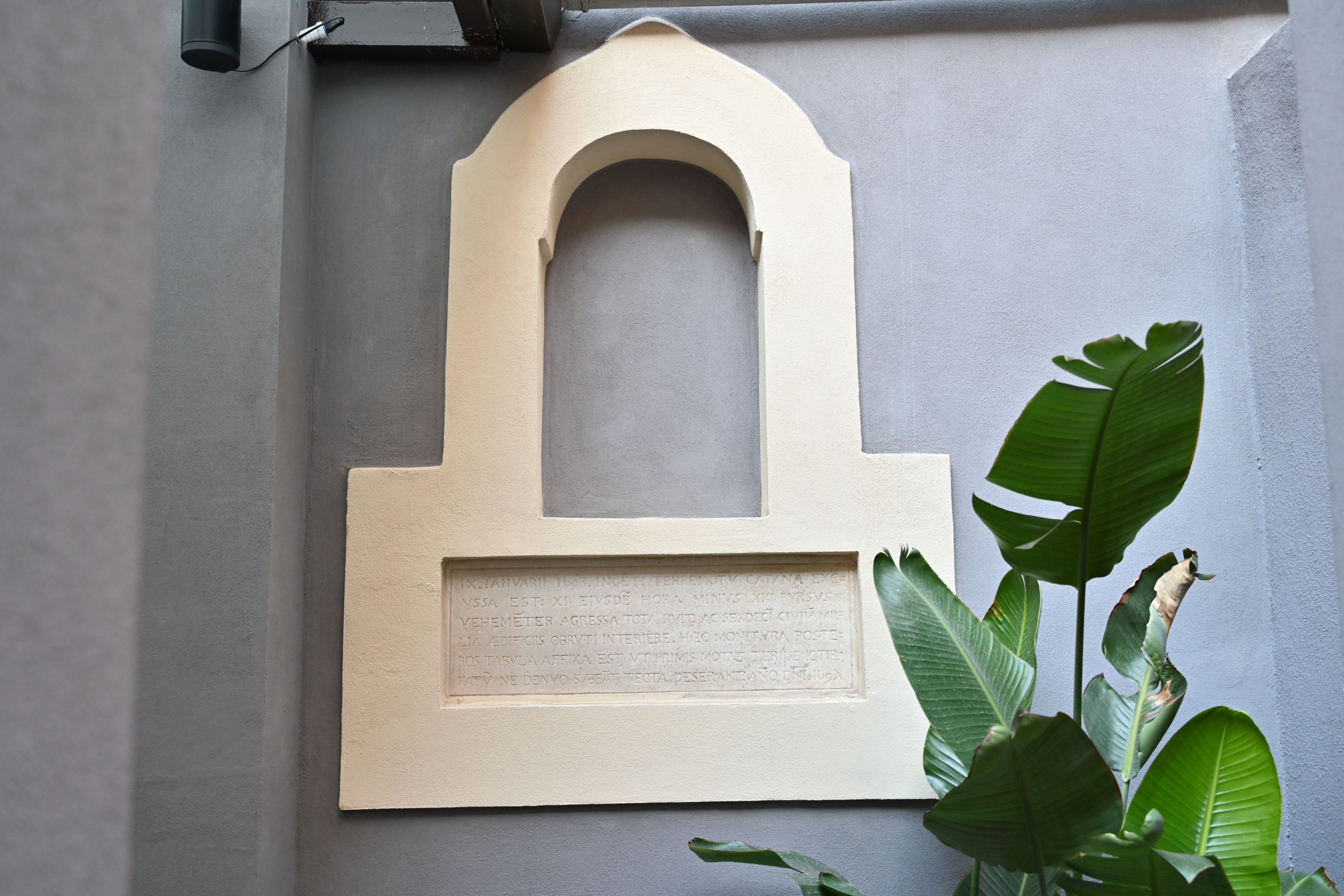 A wall-mounted plaque with an arched top and a rectangular base, surrounded by green foliage.