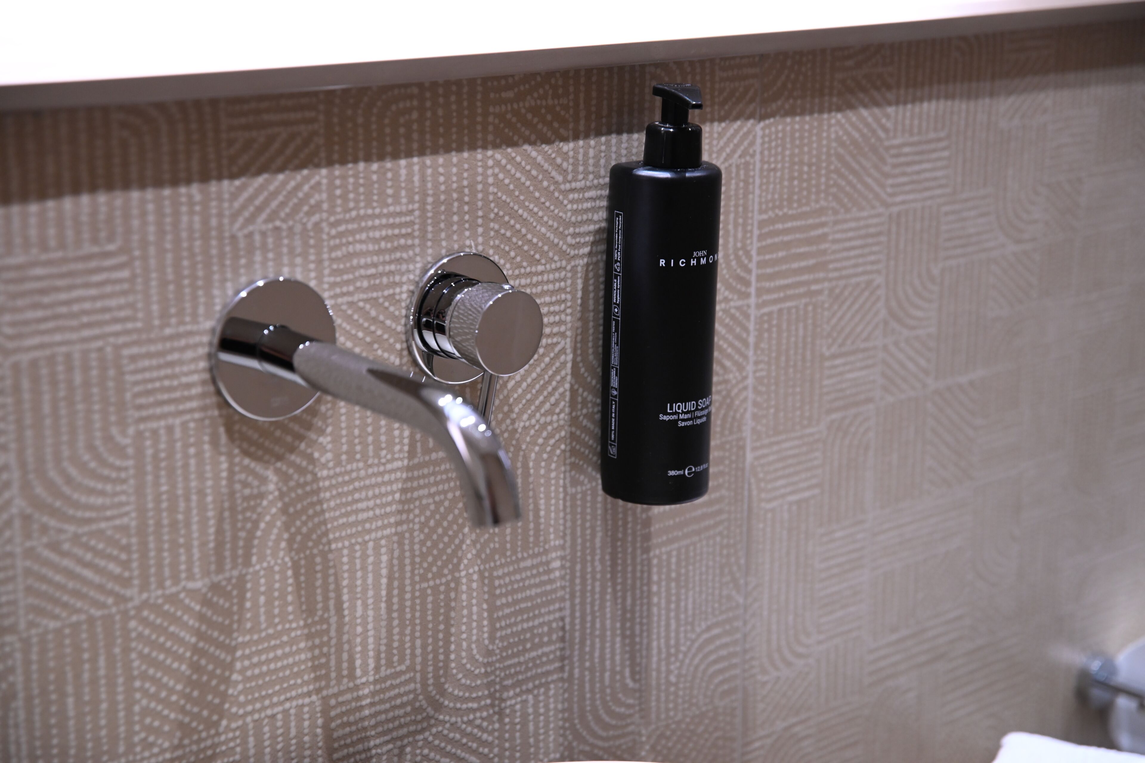 A bathroom with a patterned wall, a chrome faucet, and a black bottle of liquid soap.