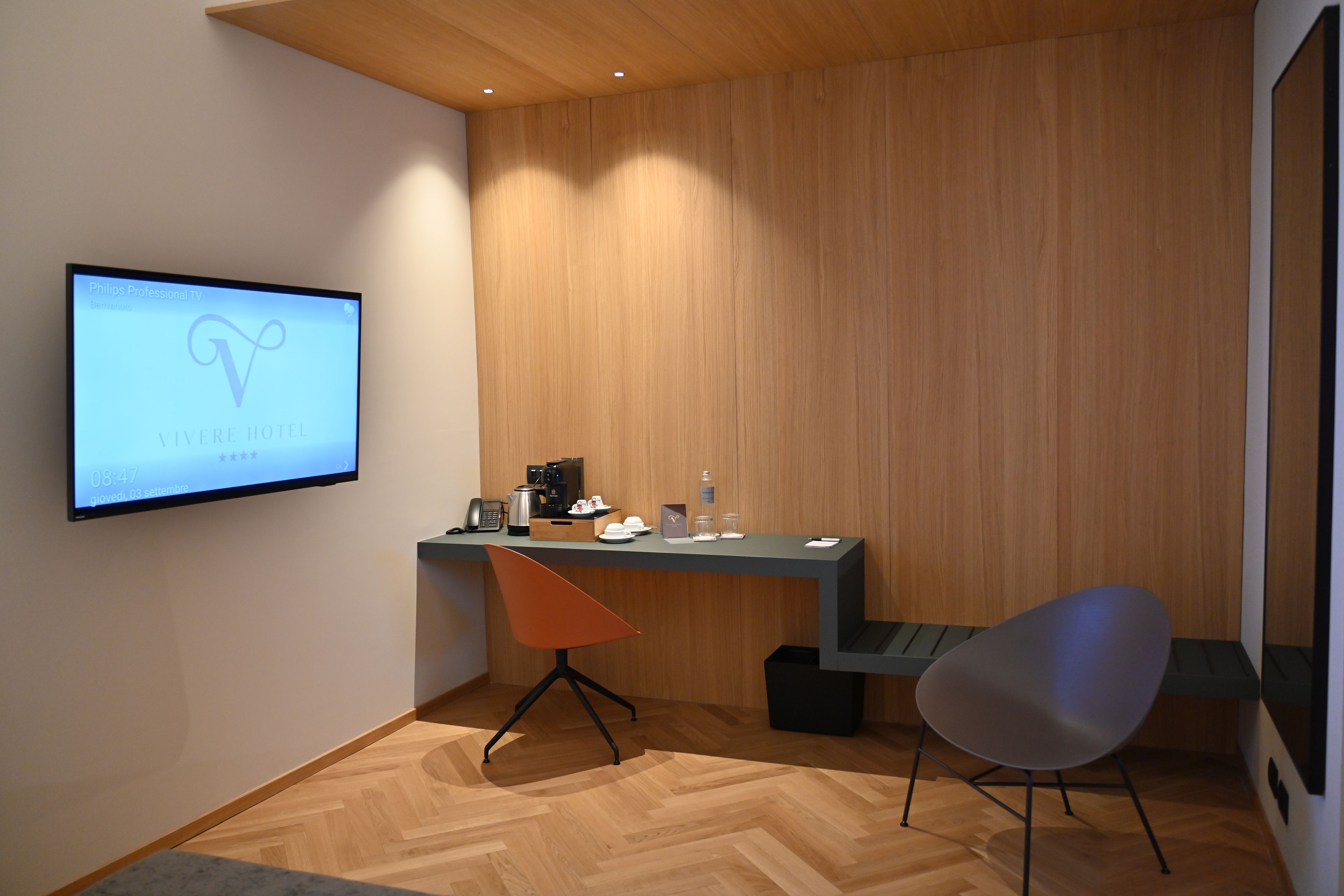 A modern hotel room with a wooden wall, a desk with a coffee maker, a chair, and a TV displaying a hotel name.