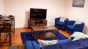 A living room with a sofa, a coffee table, a TV on a wooden stand, and a dining table with chairs.