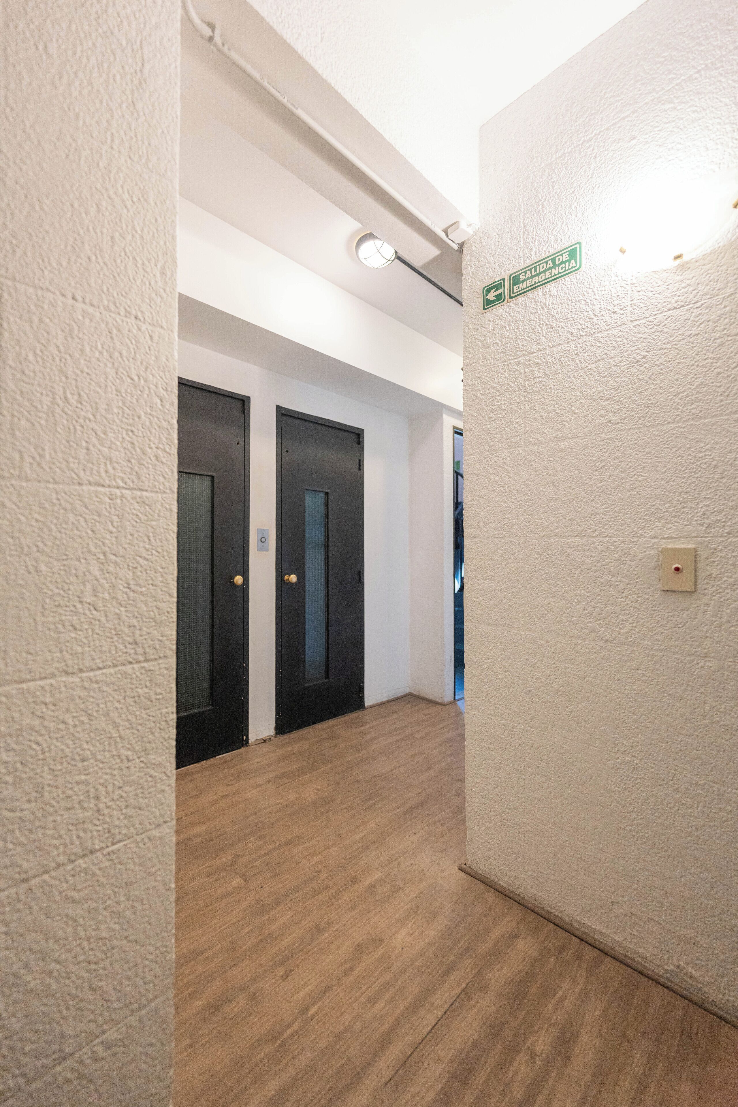 A hallway with textured walls, wooden flooring, and two black doors with a sign indicating an emergency exit.