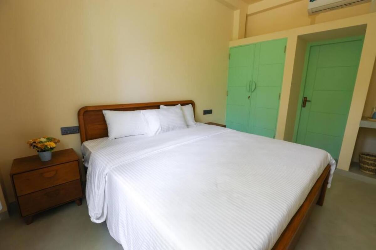 A bedroom with a wooden bed, white bedding, a nightstand with a vase of flowers, and green doors in the background.
