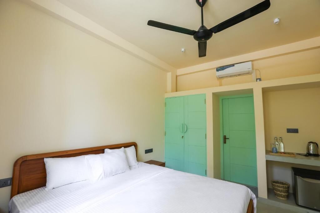 A bedroom with a bed, a ceiling fan, a green wardrobe, and a kitchenette area.