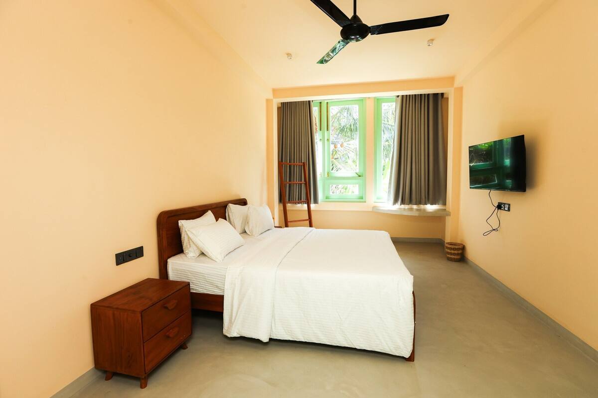 A bedroom with a bed, a nightstand, a TV, a ceiling fan, and a window with curtains.
