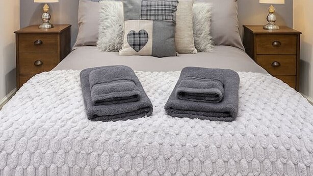 Una cama bien hecha con un edredón gris y blanco estampado, dos toallas grises dobladas y un cabecero gris.