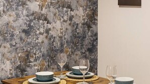 Una mesa de comedor dispuesta con platos, tazones y copas de vino, con una pared con textura y un cuadro enmarcado al fondo.