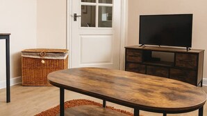 Una sala de estar con una mesa de centro de madera, un televisor sobre un mueble, una cesta de mimbre y una puerta con paneles de vidrio.