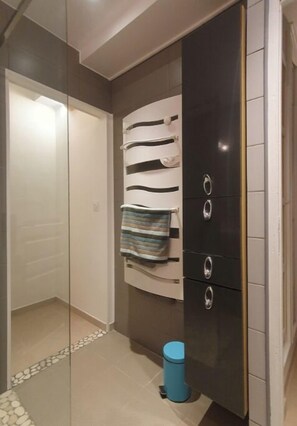 A bathroom with a glass shower enclosure, a towel rack with striped towels, a black cabinet with silver handles, and a blue trash bin. - T2 in quiet, natural villa, 5 mn from village, 20 mn from Toulon and beaches. (Le Revest-les-Eaux)