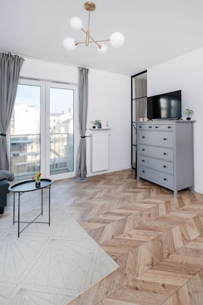 A modern living room with a herringbone wood floor, a grey cabinet with a flat-screen TV, a small table with a vase, and a large window with curtains. - Łódź Downtown - Balcony, Top Location- by Rentujemy (Łódź)