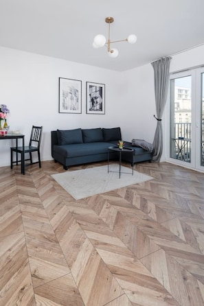 A modern living room with a herringbone wooden floor, a dark blue sofa, a small side table, and a ceiling light fixture. - Łódź Downtown - Balcony, Top Location- by Rentujemy (Łódź)