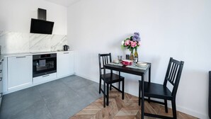 Cozinha moderna com armários brancos, forno embutido e painel de fundo em mármore. Há uma mesa de jantar com cadeiras e um vaso de flores.