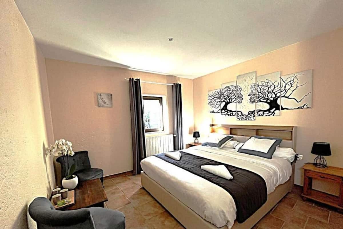 A bedroom with a bed, a chair, a small table, a lamp, and artwork on the wall.