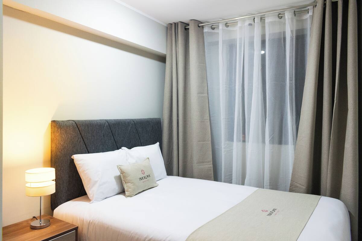 A hotel room with a bed, a nightstand with a lamp, and sheer curtains.