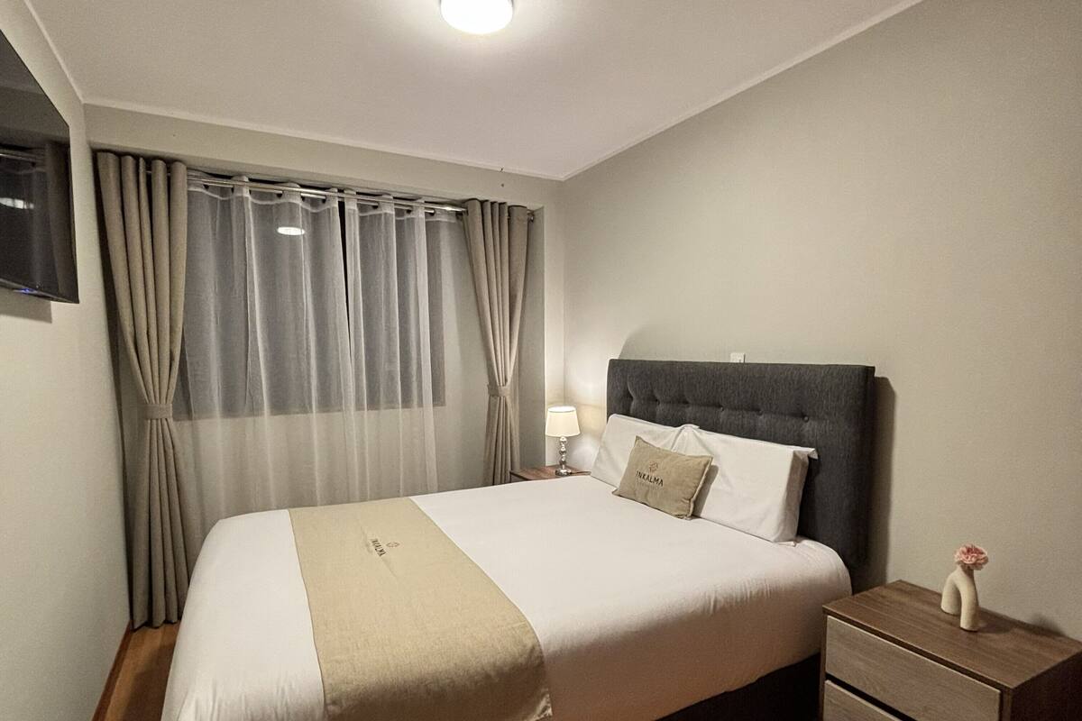 A neatly made bed with a headboard, a bedside table with a lamp, and a small nightstand with a vase.