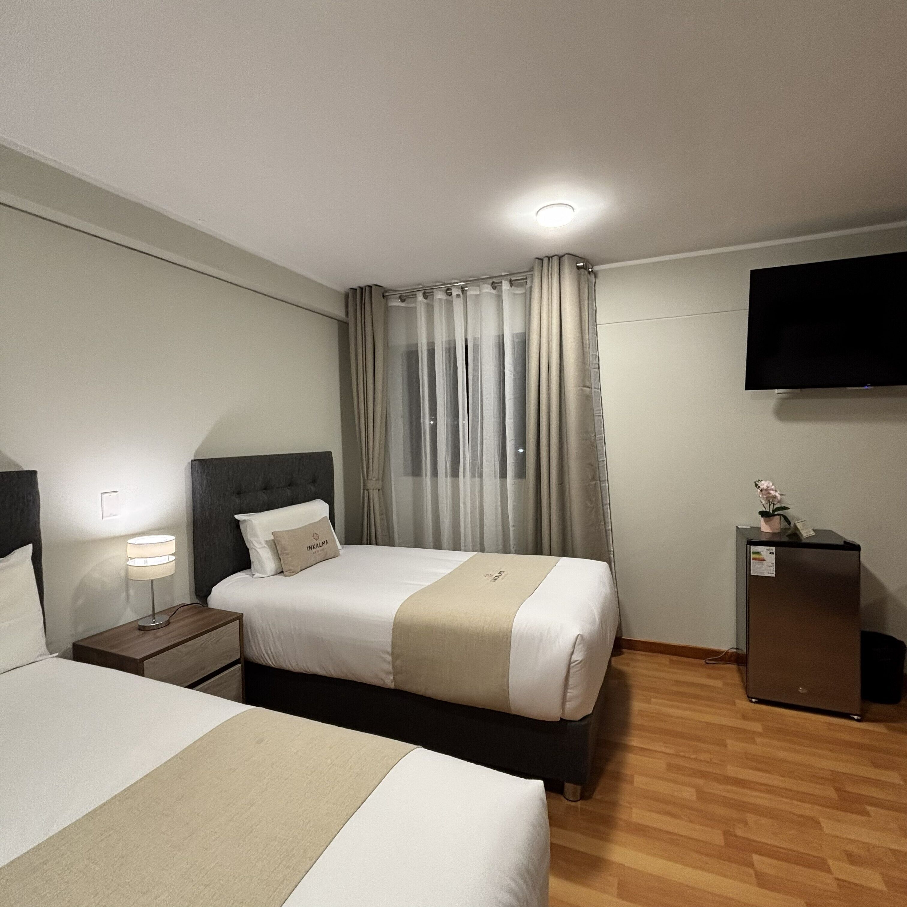 A hotel room with a bed, bedside table, lamp, curtains, a flat-screen TV, and a small refrigerator.